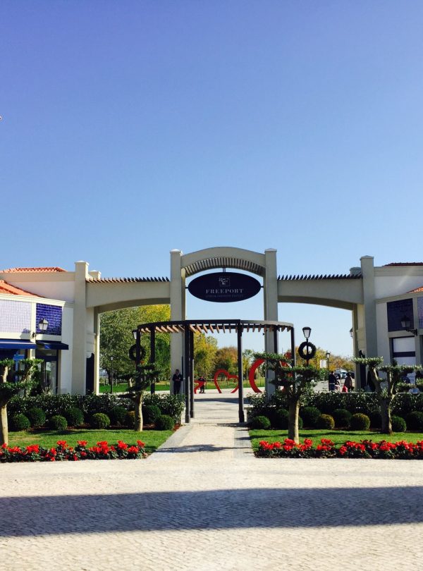 Exterior do Freeport Lisboa Fashion Outlet, com lojas ao ar livre e visitantes desfrutando de um dia de compras.