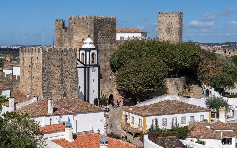 As torres do castelo e as muralhas de Óbidos sobre os telhados brancos da vila, sob um céu azul.